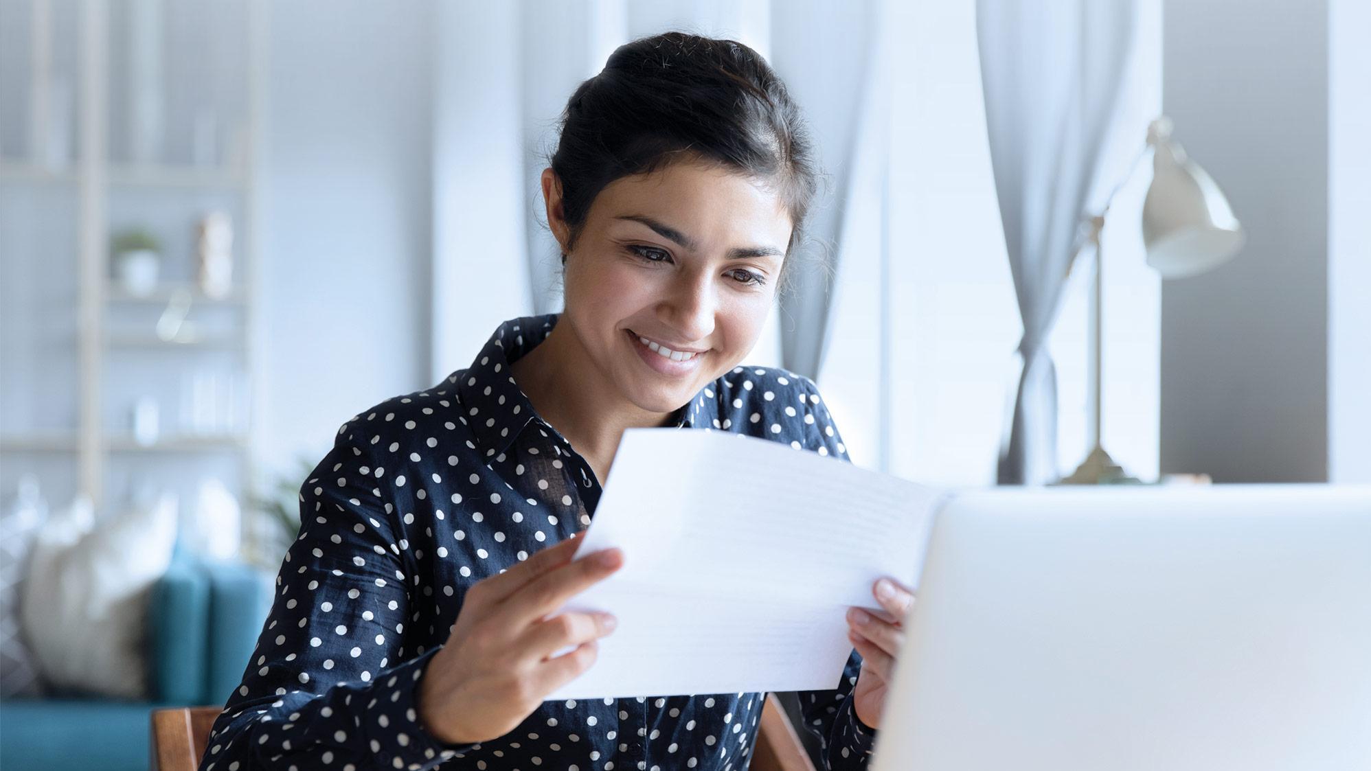 Person sitzt am Laptop und hält ein Dokument in der Hand