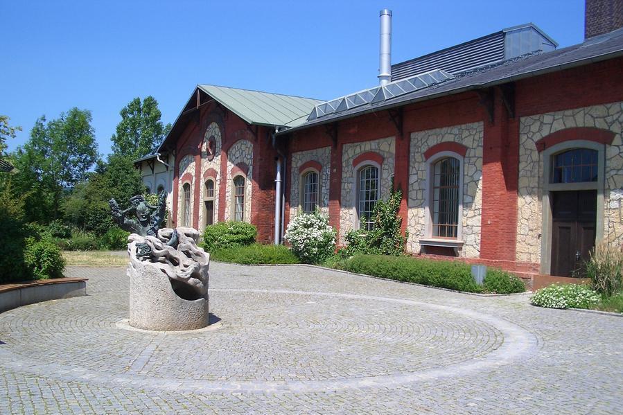 Historisches Maschinenhaus im Wasserwerk Sallern. Backsteingebäude mit Steinfassade, Innenhof und Natursteinbrunnen auf gepflastertem Platz.