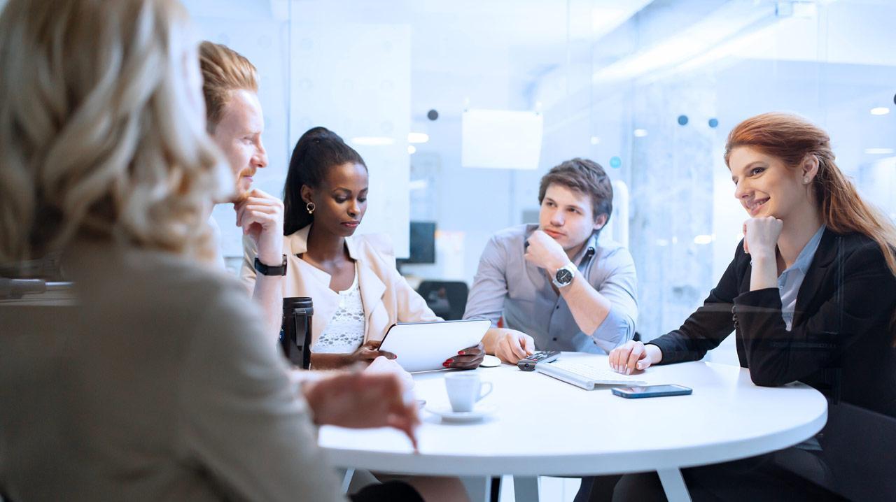 Vier Personen sitzen an einem runden Tisch in einem modernen Büro, zwei Frauen und zwei Männer, mit Dokumenten und Tassen vor sich.