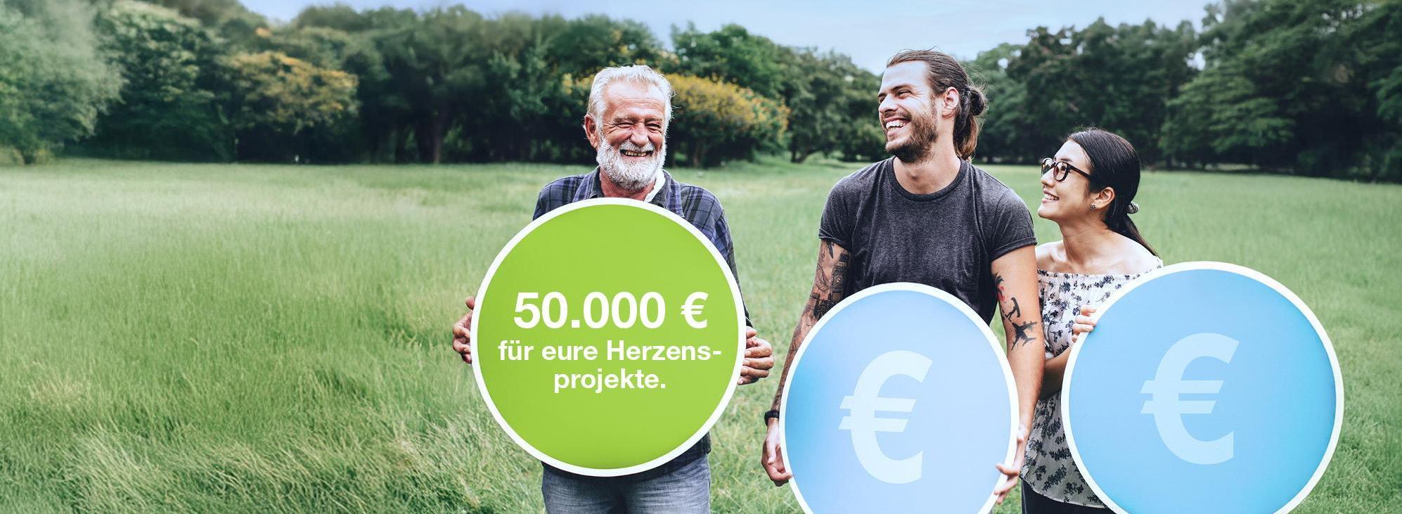 Drei Personen gehen auf einer Wiese, zwei halten große blaue Euro-Symbole, eine hält einen grünen Kreis mit dem Text '50.000 € für eure Herzensprojekte'.